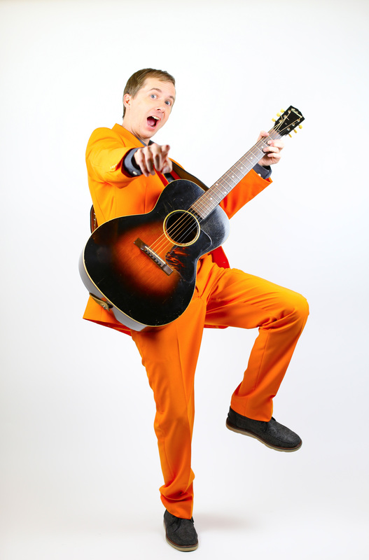 Ryan dressed in bright orange suit of The Family Fun Band smiling and holding Gibson guitar.